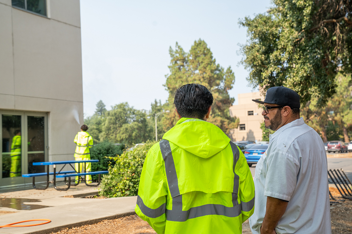 Two individuals observe as a student pressure washes the side of a campus building. One wears a neon yellow safety jacket; the other is in a white shirt and black cap. Trees, a parking lot, and a blue picnic table are visible in the background.