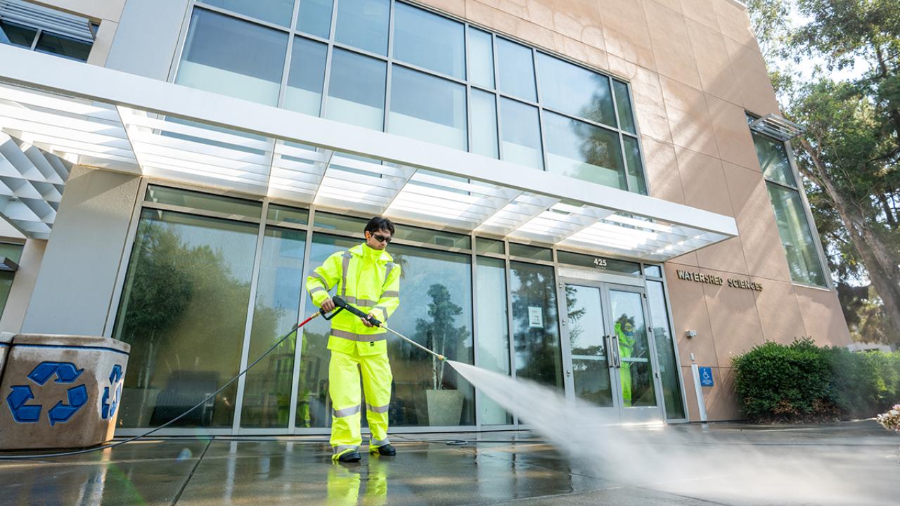 A student in a bright yellow safety suit uses a pressure washer to clean the sidewalk in front of the Watershed Sciences building at UC Davis. Water sprays outward in a strong stream as part of the summer campus refresh project.