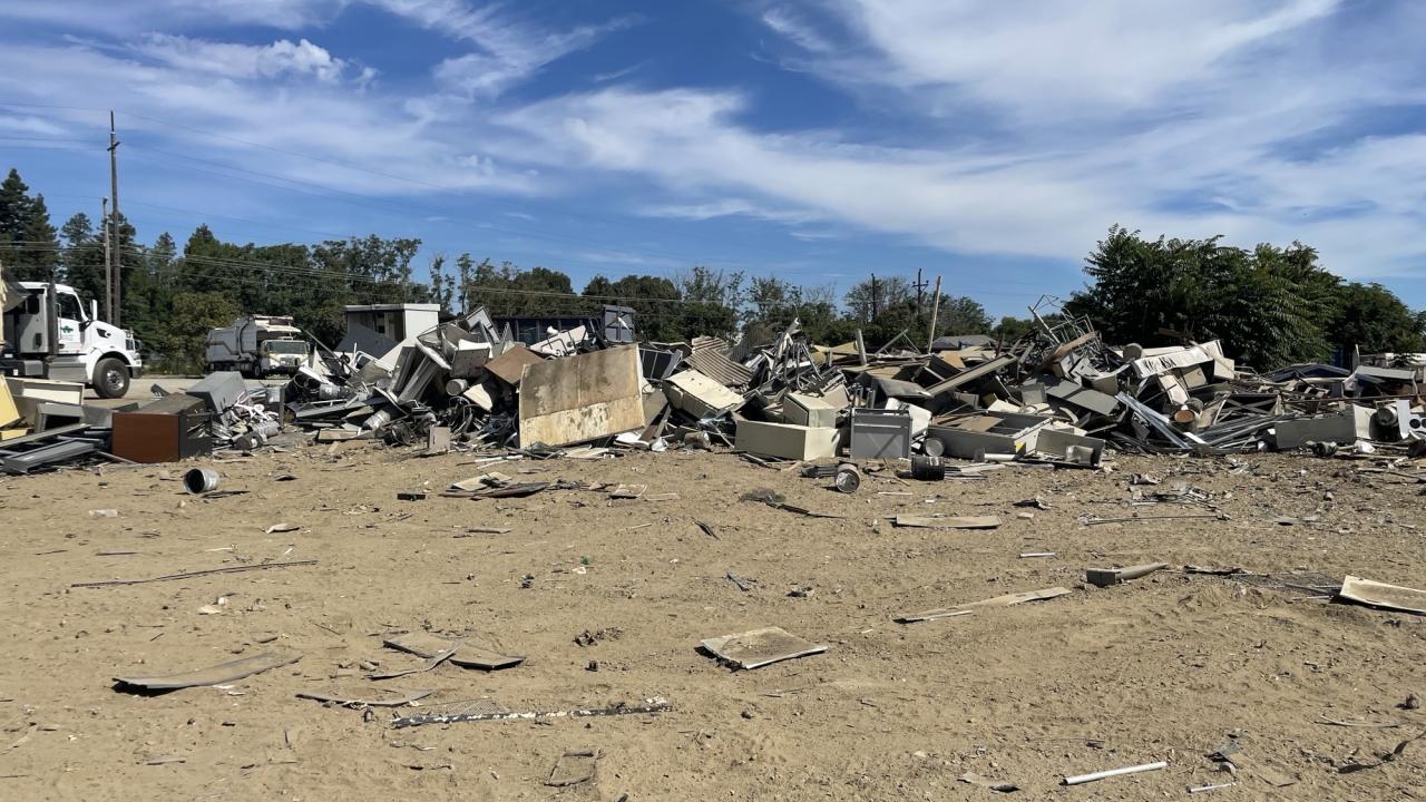 A large pile of broken furniture, metal, and construction debris scattered across a dirt lot, with two trucks parked in the background.