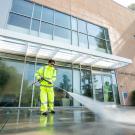 A student in a bright yellow safety suit uses a pressure washer to clean the sidewalk in front of the Watershed Sciences building at UC Davis. Water sprays outward in a strong stream as part of the summer campus refresh project.